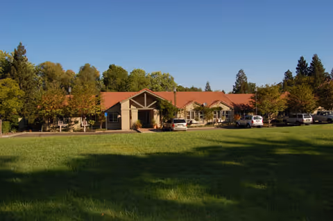 Exterior view of a single-story building with a red-tiled roof surrounded by trees and a green lawn in the foreground under a clear blue sky. Several cars are parked near the building.