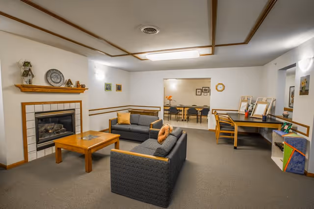 A cozy living room area with two dark gray sofas facing each other around a wooden coffee table. There is a fireplace with a wooden mantel decorated with small items and a clock on the wall. The room has beige walls with wooden trim and carpeted flooring. In the background, there is a doorway leading to another room with a table and chairs. On the right side, there is a wooden desk with chairs and some art supplies.