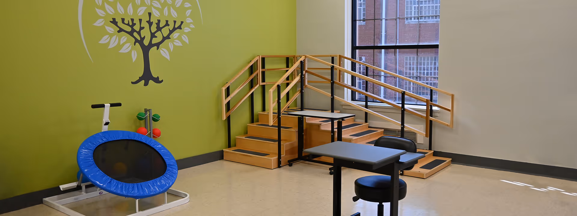 Rehabilitation therapy room with parallel bars and exercise stairs, a small blue trampoline and seating against a green wall with a tree decal and a window.