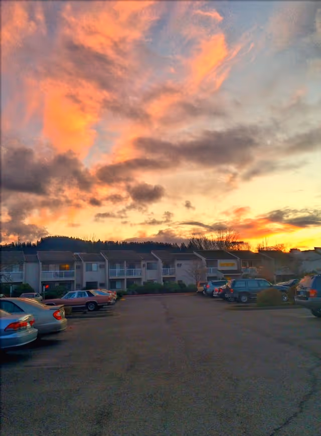 Parking lot in front of a two-story residential building with balconies under a colorful sunset sky with orange and pink clouds.