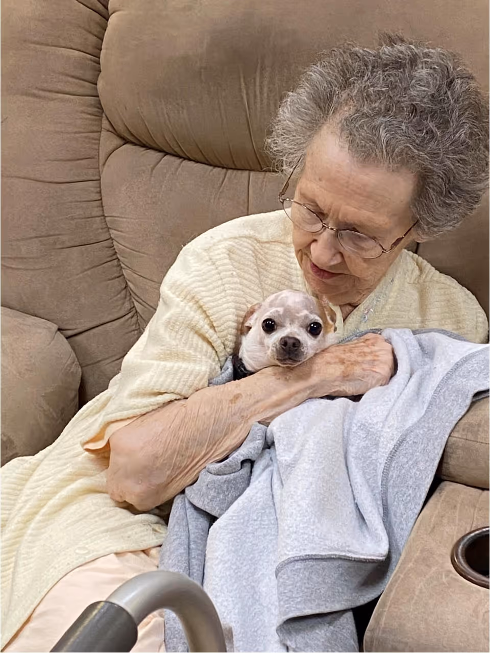 An elderly woman with glasses sitting on a beige recliner chair, gently holding a small dog wrapped in a gray blanket.