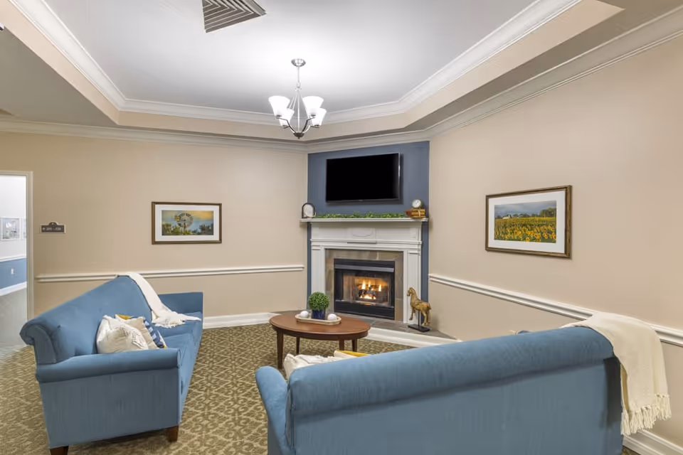 A cozy living room with two blue sofas facing a fireplace with a mounted flat-screen TV above it. The room has beige walls with white trim, a patterned carpet, a round wooden coffee table with decorative items, and framed landscape paintings on the walls. A ceiling light fixture hangs above.