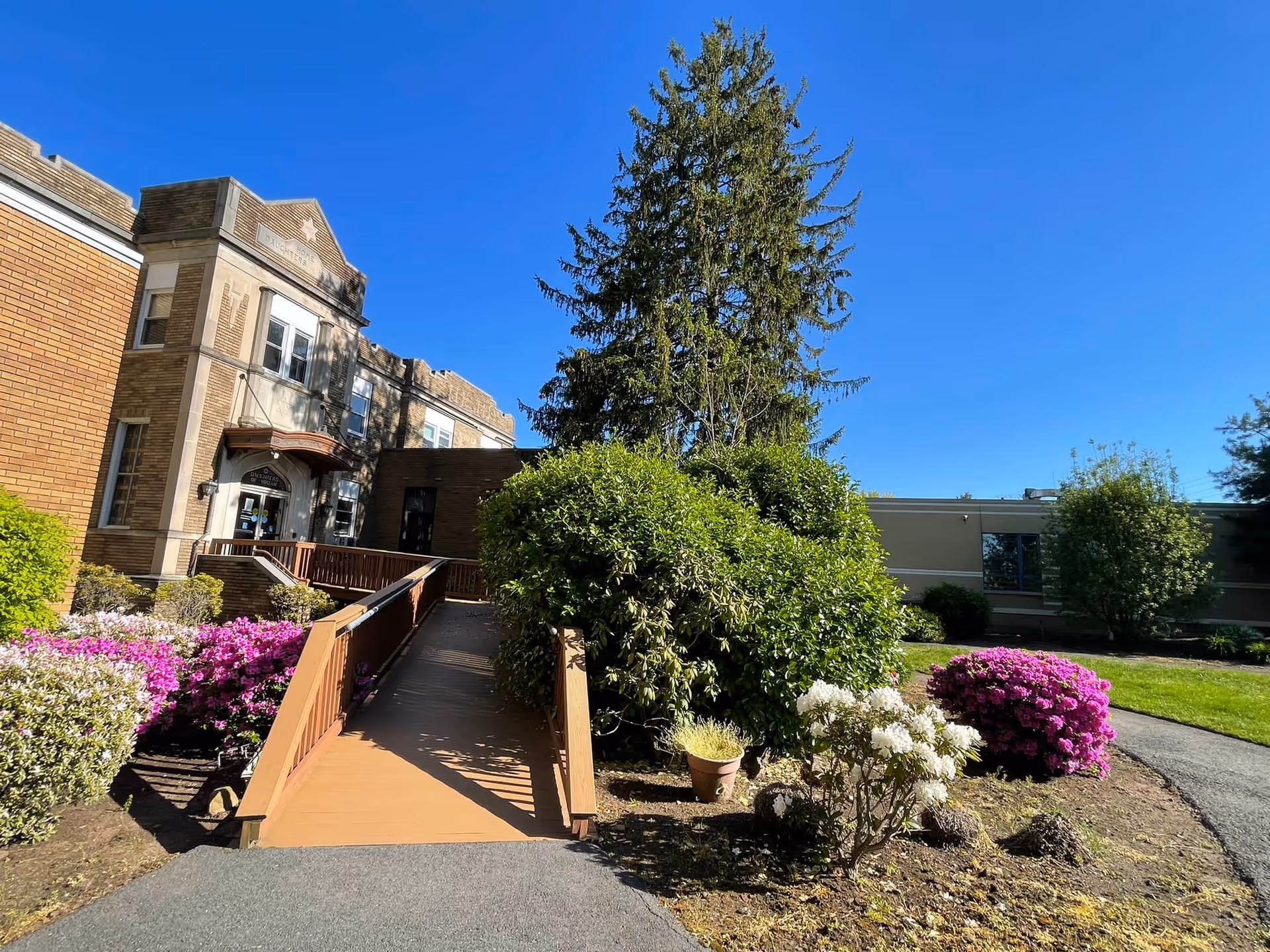 A wooden ramp leads to the entrance of a brick institutional building surrounded by flowering shrubs and a large evergreen under a clear blue sky.