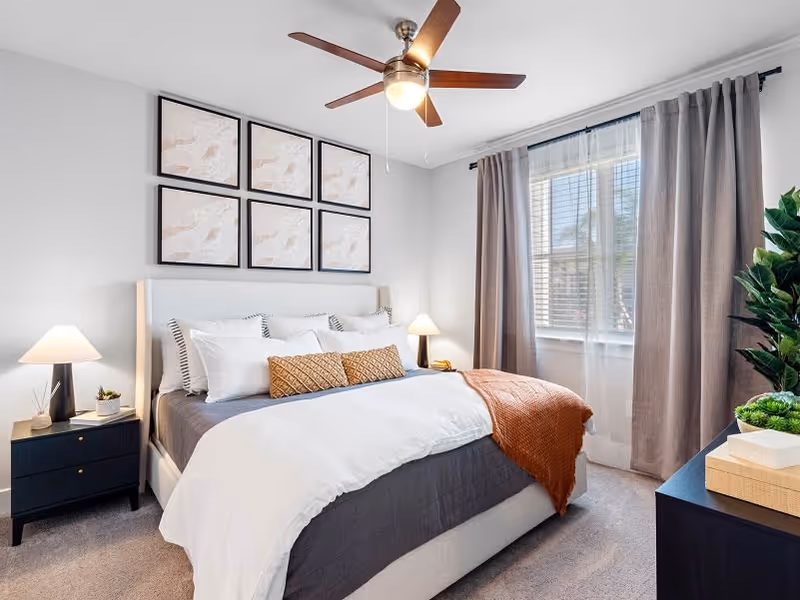 A bright and cozy bedroom featuring a large bed with white and gray bedding, decorative pillows, and an orange throw blanket. The room has a ceiling fan with a light, two bedside tables with lamps, a window with gray curtains, and framed artwork on the wall above the bed. There is also a plant and decorative items on a dresser.