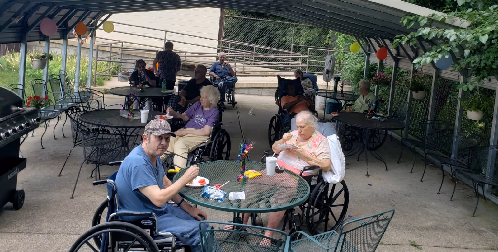Several elderly individuals, some in wheelchairs, are seated around metal tables under a covered outdoor patio area. They appear to be eating and socializing. The patio is decorated with hanging flower pots and colorful balloons. There is a grill on the left side of the image and greenery surrounding the area.