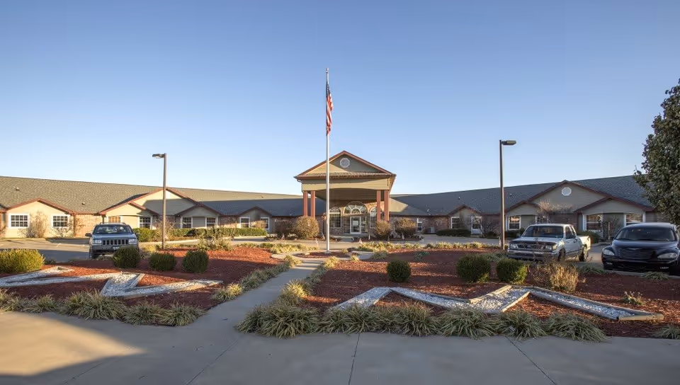 Exterior view of Emerald Care Center Claremore building with a central entrance featuring a covered porch and an American flagpole in front. The landscaped area includes decorative mulch, bushes, and plants, with several parked vehicles visible on either side of the entrance.