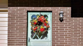 A mint-green front door set in a brick wall, decorated with a large floral wreath and an exterior light fixture.