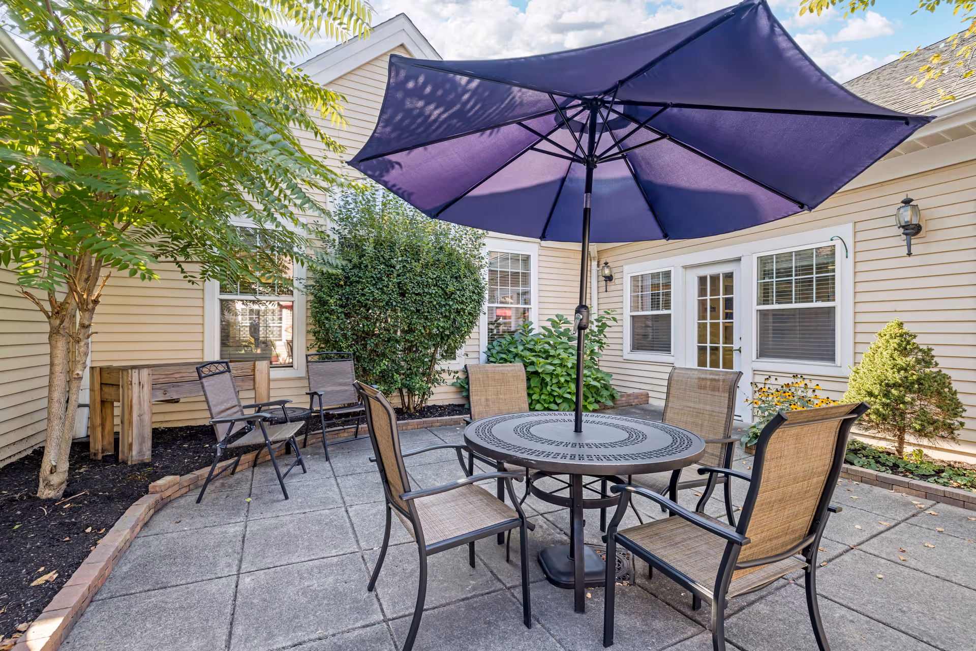 Outdoor patio area with a round metal table and four chairs under a large purple umbrella. The patio is surrounded by beige siding walls of a building, with windows and a glass door. There are green bushes, a small tree, and other plants around the patio, along with a wooden bench and additional chairs.