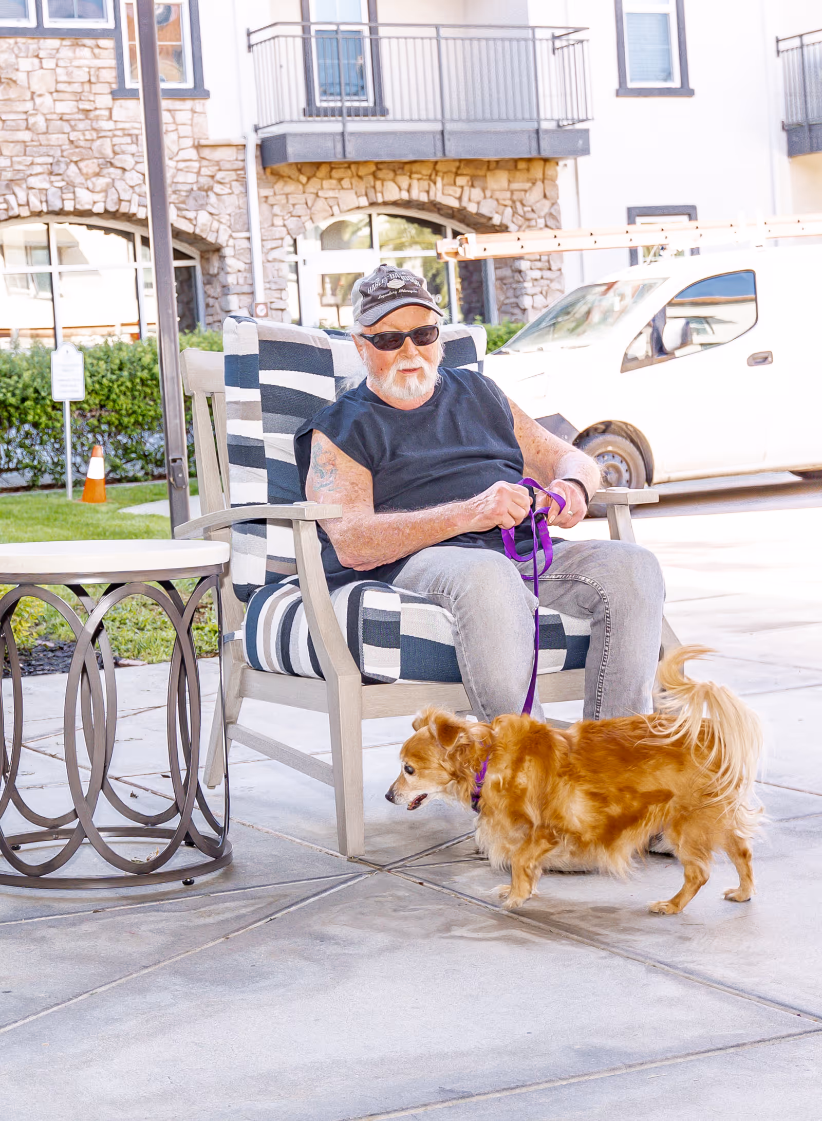 An elderly man wearing sunglasses, a black sleeveless shirt, and a cap is sitting on a cushioned outdoor chair holding a purple leash attached to a small brown dog. They are on a paved patio area outside a building with stone and stucco exterior walls. A white van is parked in the background.