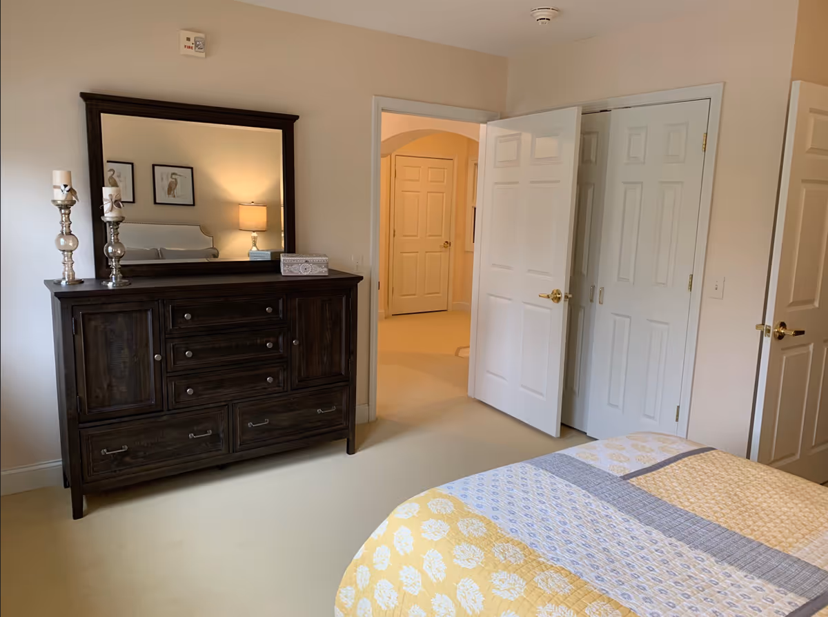 Well-lit bedroom showing a dark wooden dresser with a large mirror and decorative candles, an open door to a hallway, and part of a bed with a yellow quilt.