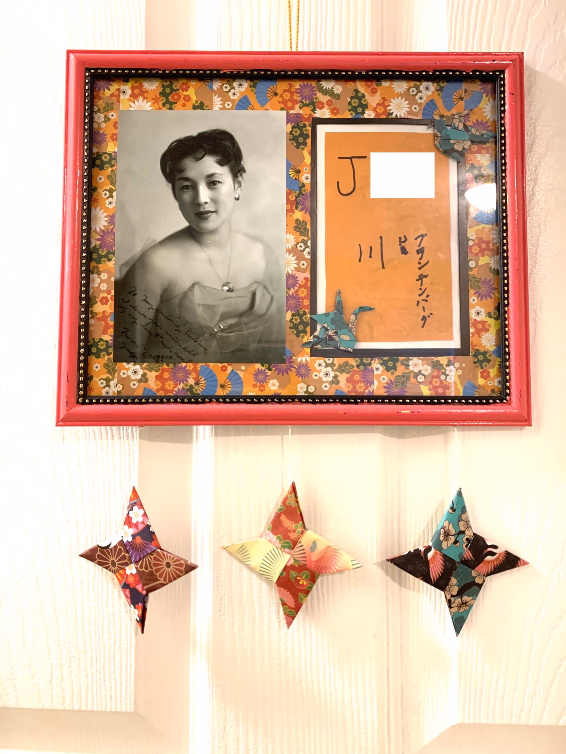 A framed vintage portrait and a handwritten card with floral matting hung on a white door above three colorful origami star decorations.