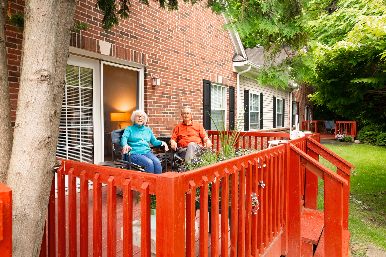 An elderly woman and man sitting on chairs on a red wooden porch outside a brick and siding building. The porch has a railing with potted plants, and there is greenery and trees surrounding the area.