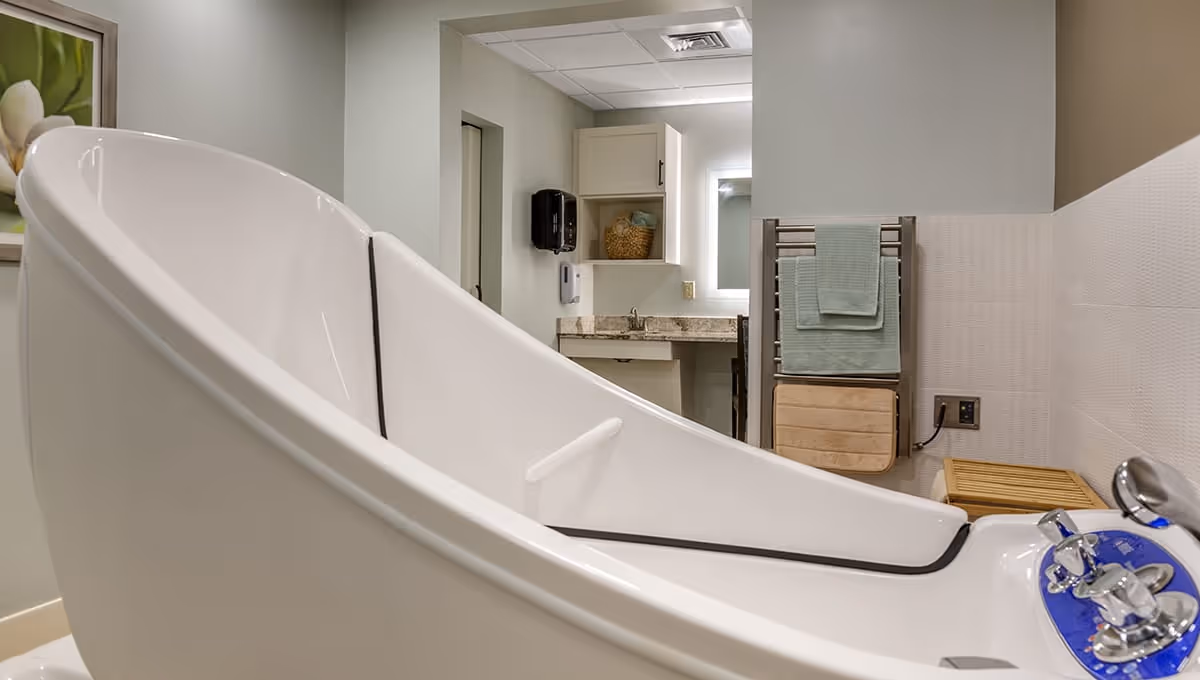 A modern bathroom featuring a large white therapeutic bathtub with control knobs, a towel rack with folded towels, a wooden bench, a countertop with a sink, and a wall-mounted soap dispenser. The walls are painted light green and white, and there is a framed floral picture on the wall.