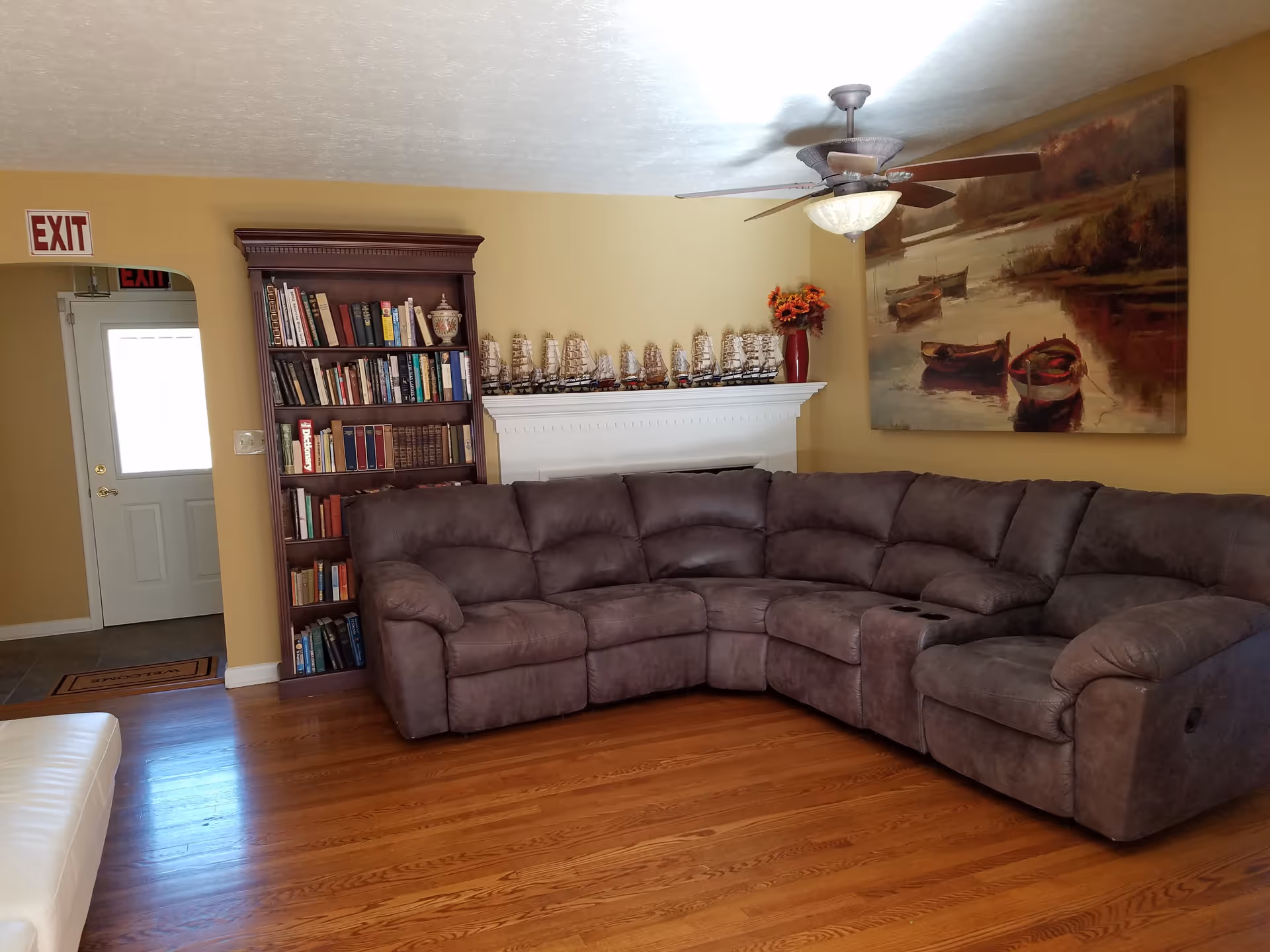 A cozy living room with a large brown sectional sofa, a wooden bookshelf filled with books, and a white mantel decorated with multiple model ships and a vase of flowers. A ceiling fan with a light hangs above, and a large painting of boats on water is mounted on the wall. The room has wooden flooring and yellow walls, with an exit door visible in the background.