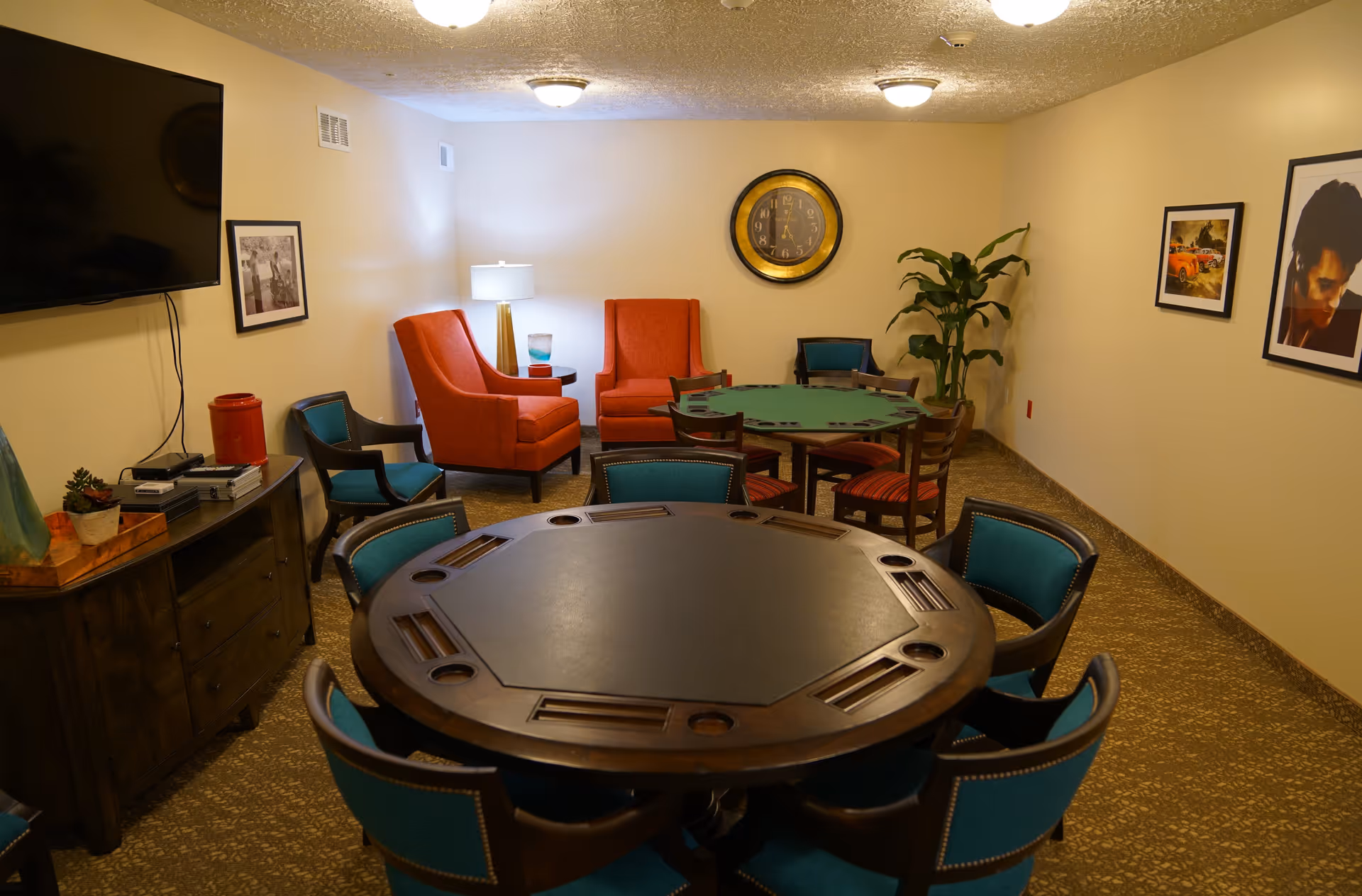 A cozy game room with two round poker tables surrounded by chairs, two orange armchairs near a side table with a lamp, a large wall clock, framed pictures on the walls, a flat-screen TV mounted on the wall, and a wooden cabinet with decorative items.