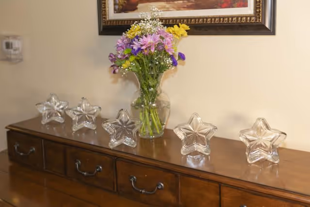 A wooden dresser with five clear star-shaped glass ornaments arranged in a row on top. In the center, there is a glass vase holding a bouquet of colorful flowers including purple, yellow, and white blooms. A framed painting hangs on the beige wall above the dresser.