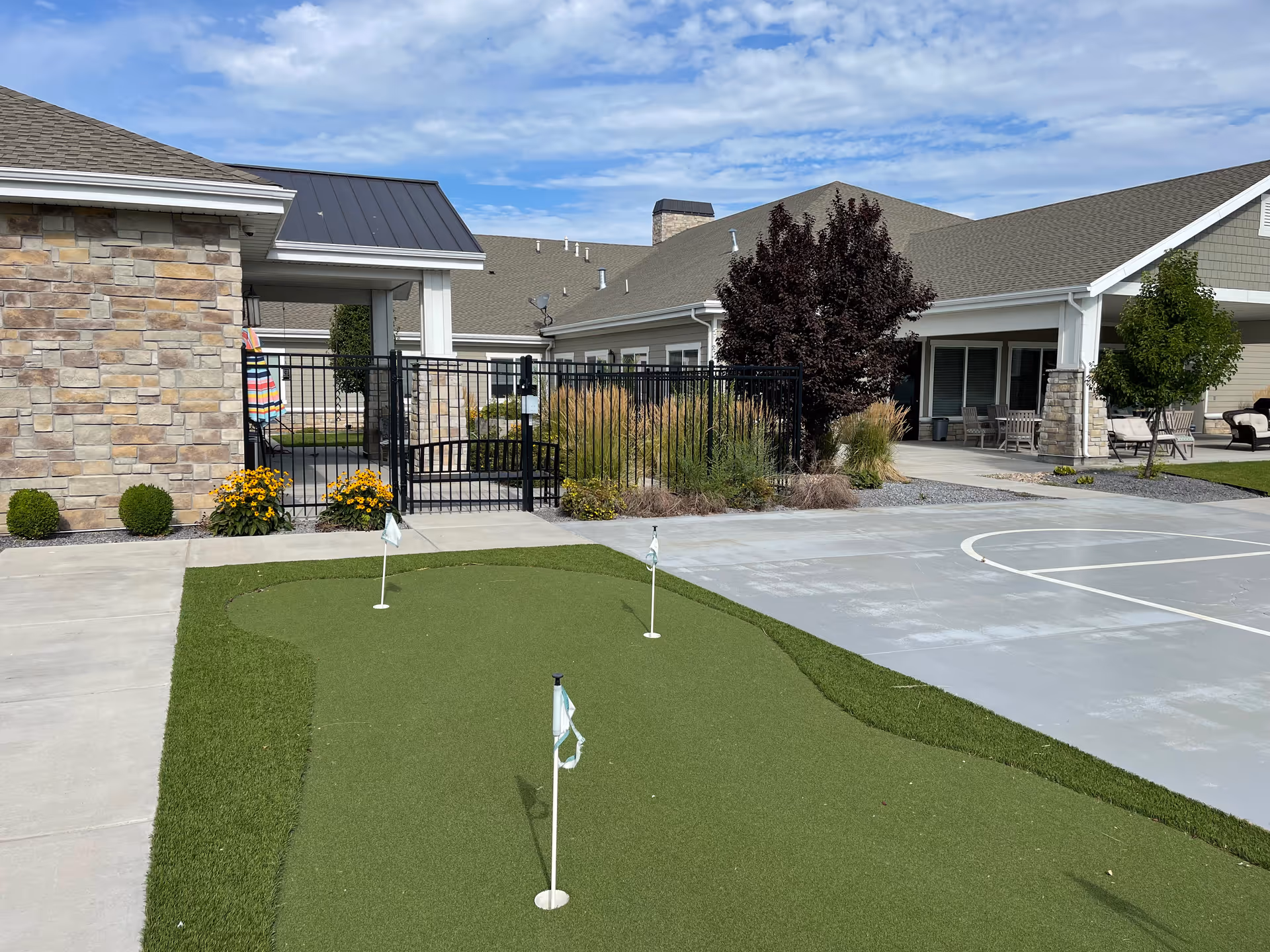 Outdoor area of Sunridge Assisted Living of Layton featuring a small artificial putting green with three flags, a basketball court, stone and siding building exterior, patio seating, and landscaping with bushes and trees under a partly cloudy sky.