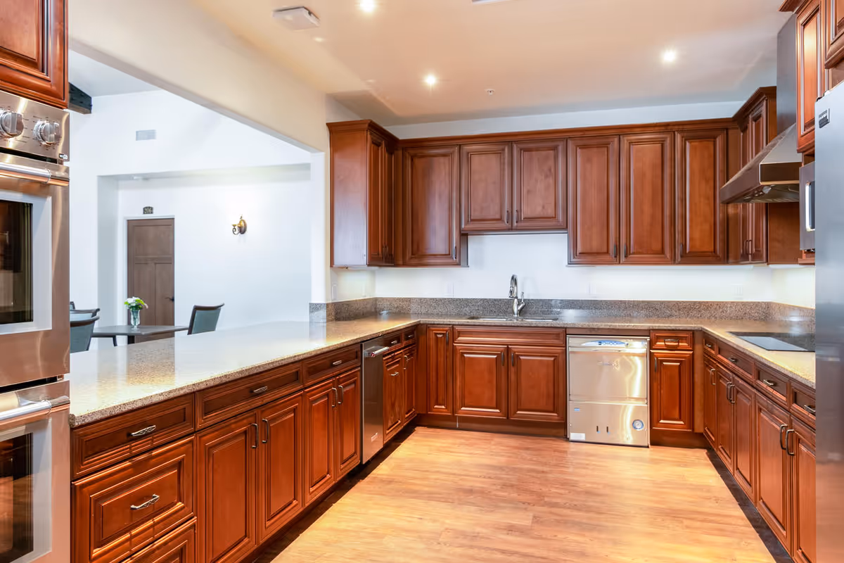 A spacious kitchen with wooden cabinets, granite countertops, stainless steel appliances including a double oven, dishwasher, and refrigerator. The kitchen has a wooden floor and recessed lighting. In the background, there is a dining area with a table and chairs and a wall sconce light.