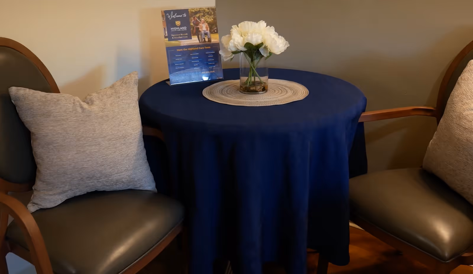 A small round table covered with a dark blue tablecloth, with a vase of white flowers and a welcome sign for Highland Healthcare Center on top. Two chairs with cushions are positioned on either side of the table against a beige wall.