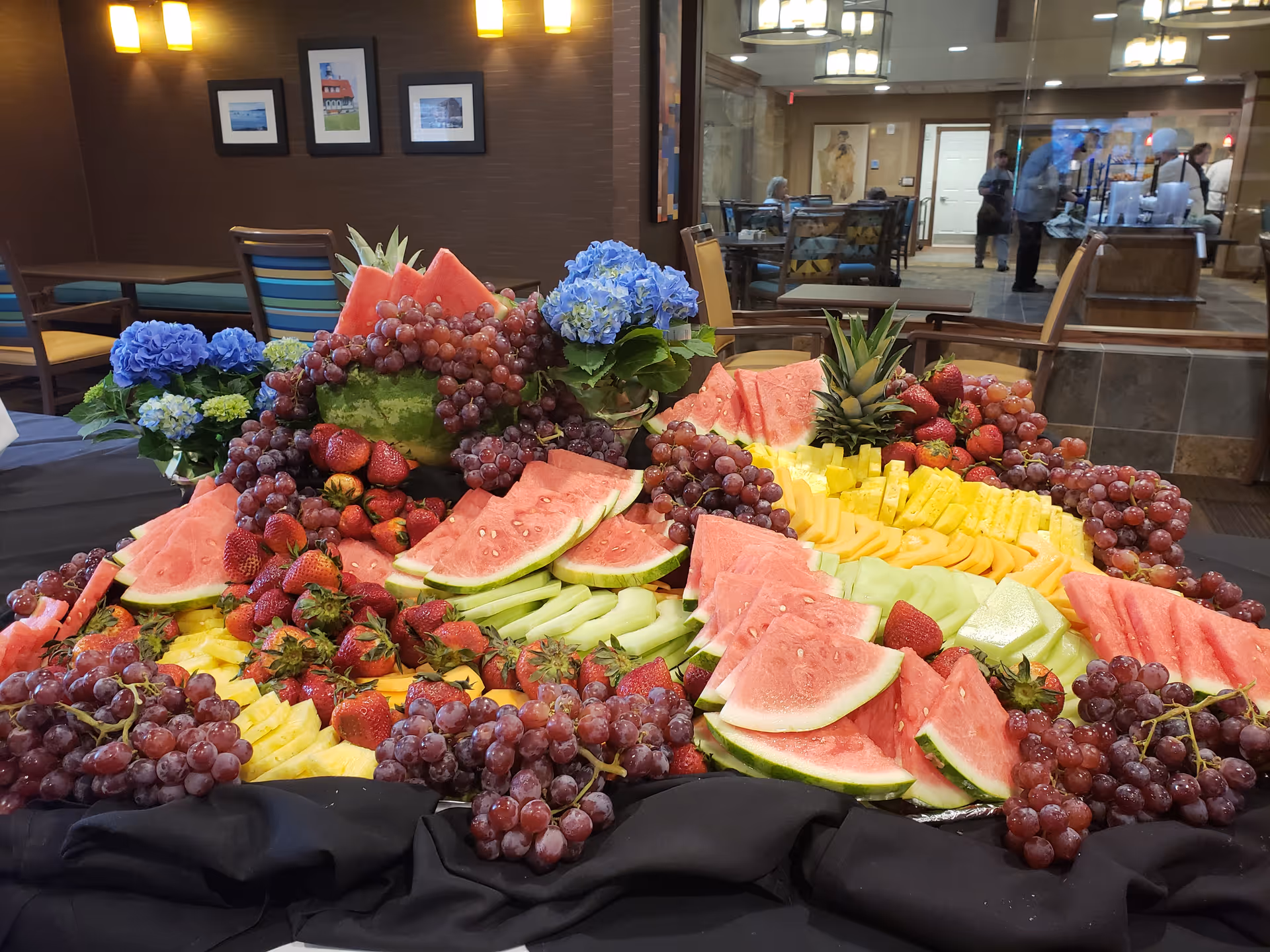 A large, colorful fruit platter featuring sliced watermelon, grapes, strawberries, pineapple, cantaloupe, and honeydew melon arranged on a black tablecloth. Blue hydrangea flowers are placed around the platter. In the background, there is a dining area with tables, chairs, and people visible through a glass partition.