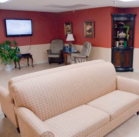 A cozy communal living room with patterned sofas, armchairs, a wall-mounted TV, lamp, and a corner shelving unit.