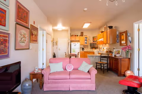Interior view of a senior living facility showing a cozy living room with a pink sofa adorned with two light green pillows and a round decorative cushion. The room includes a wooden side table with a lamp, a TV on a stand, and several framed artworks on the wall. In the background, there is a kitchen area with wooden cabinets, a refrigerator, and a dining table with chairs. The space is warmly lit and decorated with flowers and various household items.