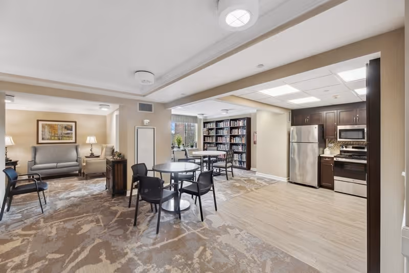A well-lit common area in a senior living facility featuring a small kitchen with stainless steel appliances including a refrigerator, stove, and microwave. The room has round tables with black chairs, a seating area with a sofa and armchairs, and a bookshelf filled with books near a window.
