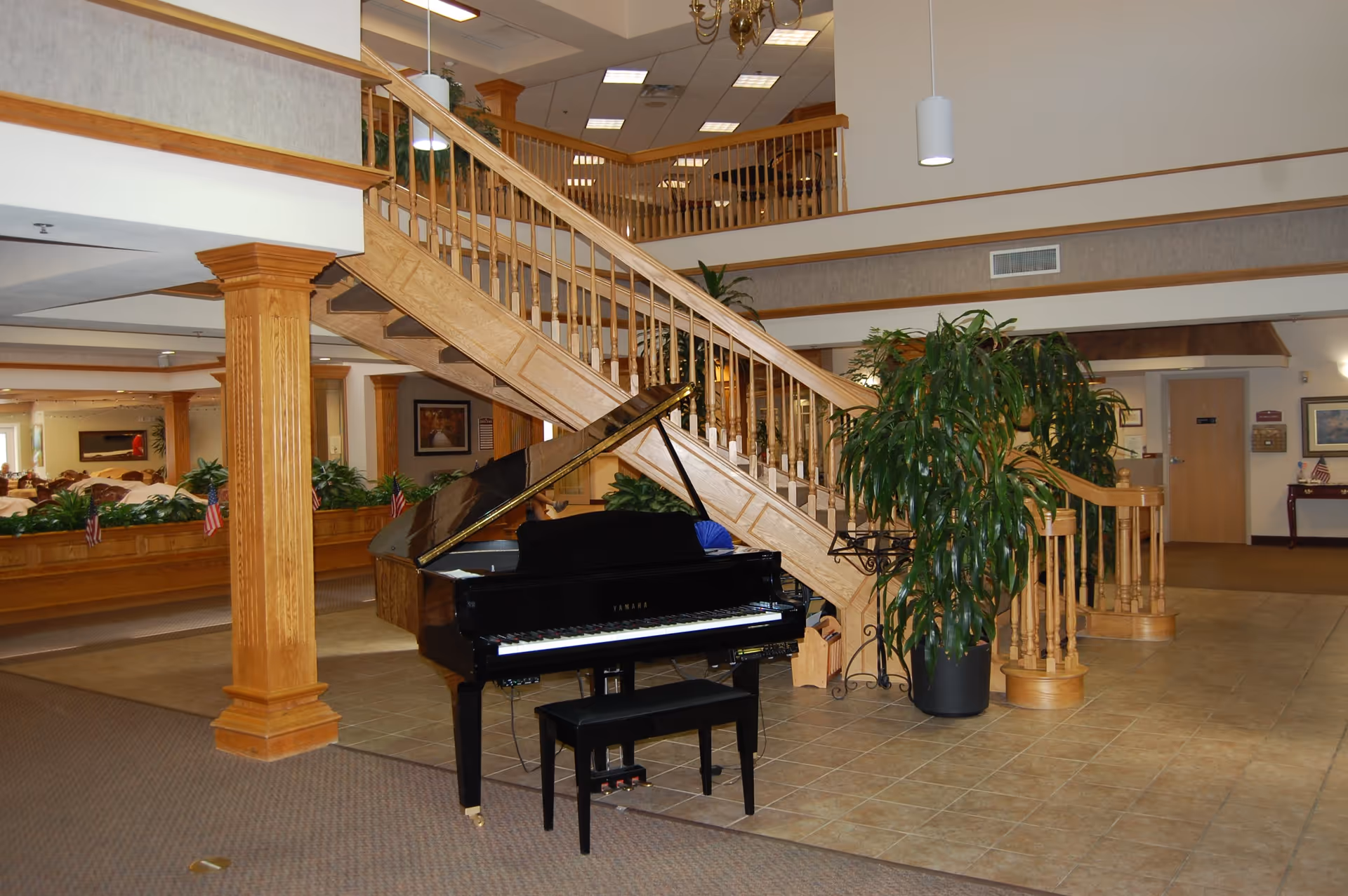 Interior view of an assisted living community featuring a black grand piano with a bench in the foreground, a wooden staircase with railings leading to an upper floor, large potted plants, and a dining area visible in the background with tables and chairs.
