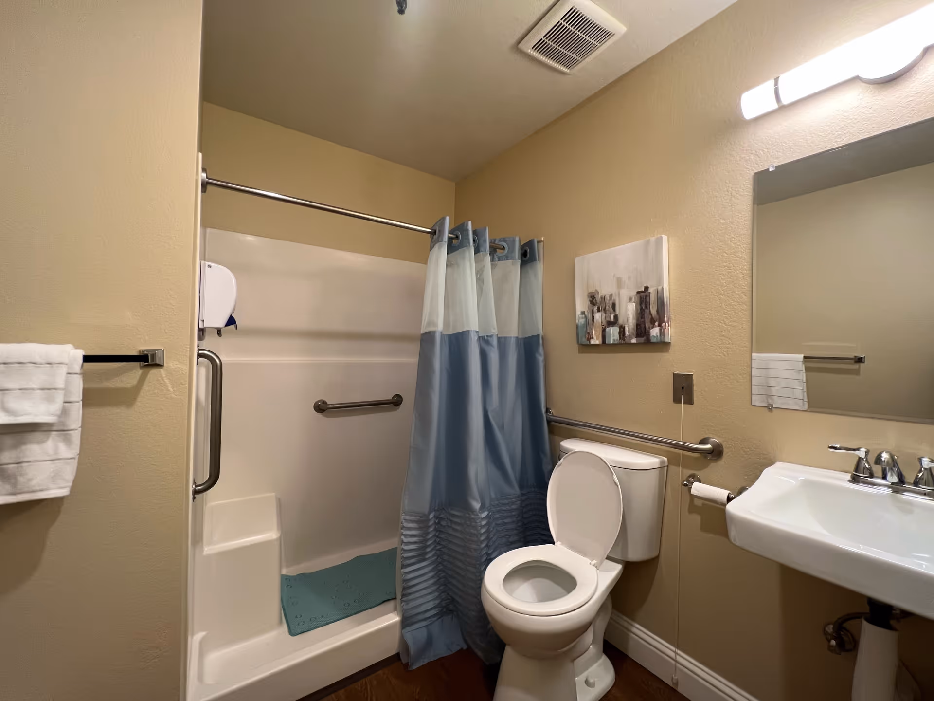 A bathroom with beige walls featuring a white toilet with the lid open, a white sink with a silver faucet, a large mirror above the sink, and a shower with a blue and white curtain. There are metal grab bars near the toilet and inside the shower, a towel hanging on a rack, a blue bath mat inside the shower, and a small piece of artwork on the wall.