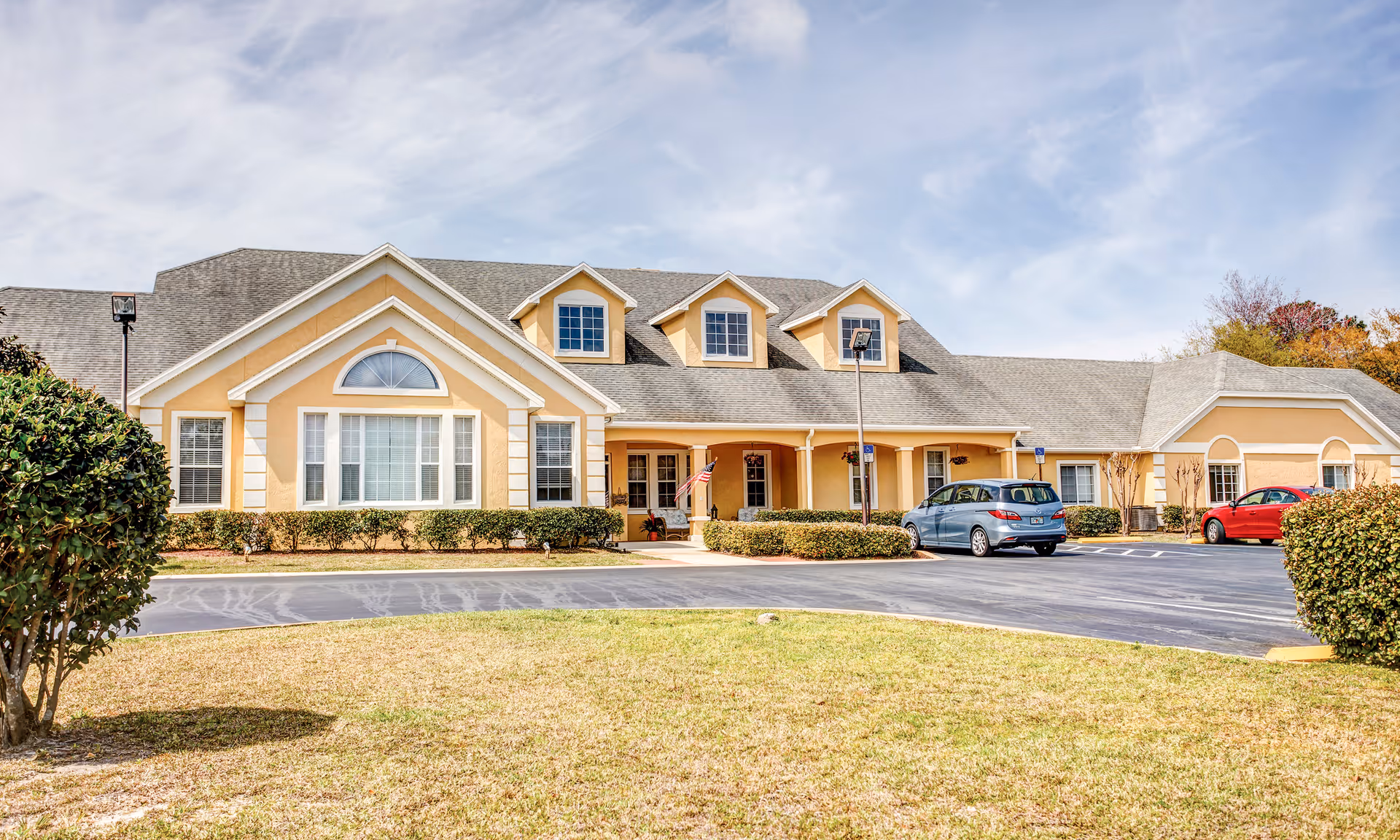 Exterior front view of a single-story senior living facility building with a beige facade, multiple windows, and a gray shingled roof. There are three dormer windows on the roof and a covered entrance with an American flag. Two cars are parked in the parking lot in front of the building, and there are bushes and a grassy area in the foreground under a partly cloudy sky.