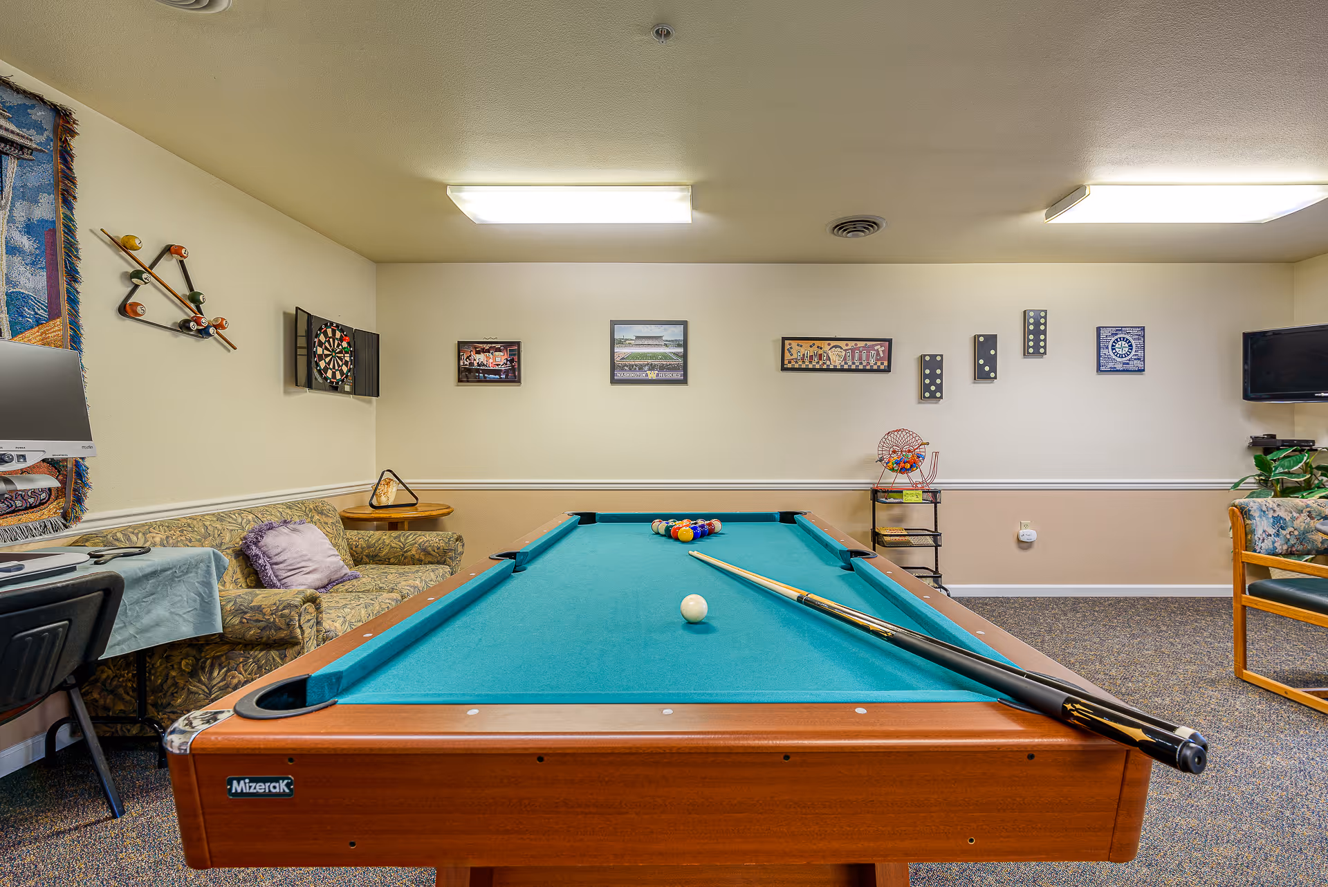 Pool table in a recreation room with sofas, a dartboard, wall decor, and a TV.