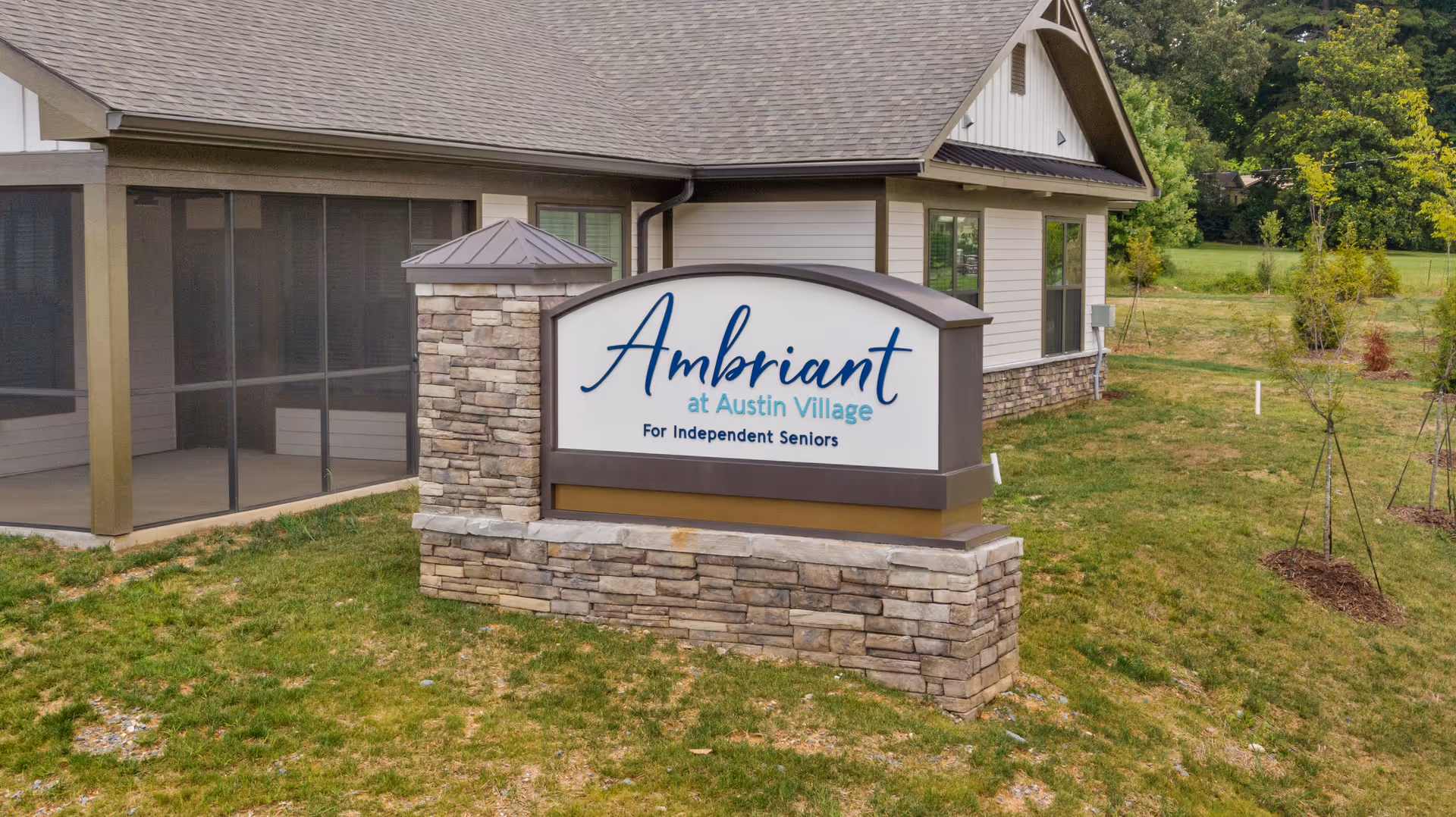 Outdoor view of a senior living facility sign that reads 'Ambriant at Austin Village For Independent Seniors' in front of a building with stone and siding exterior and a screened porch, surrounded by grass and small trees.