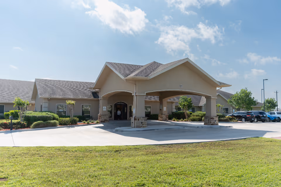 Front exterior of a single-story senior living facility with a covered porte-cochere, landscaped grounds and parked cars under a blue sky.