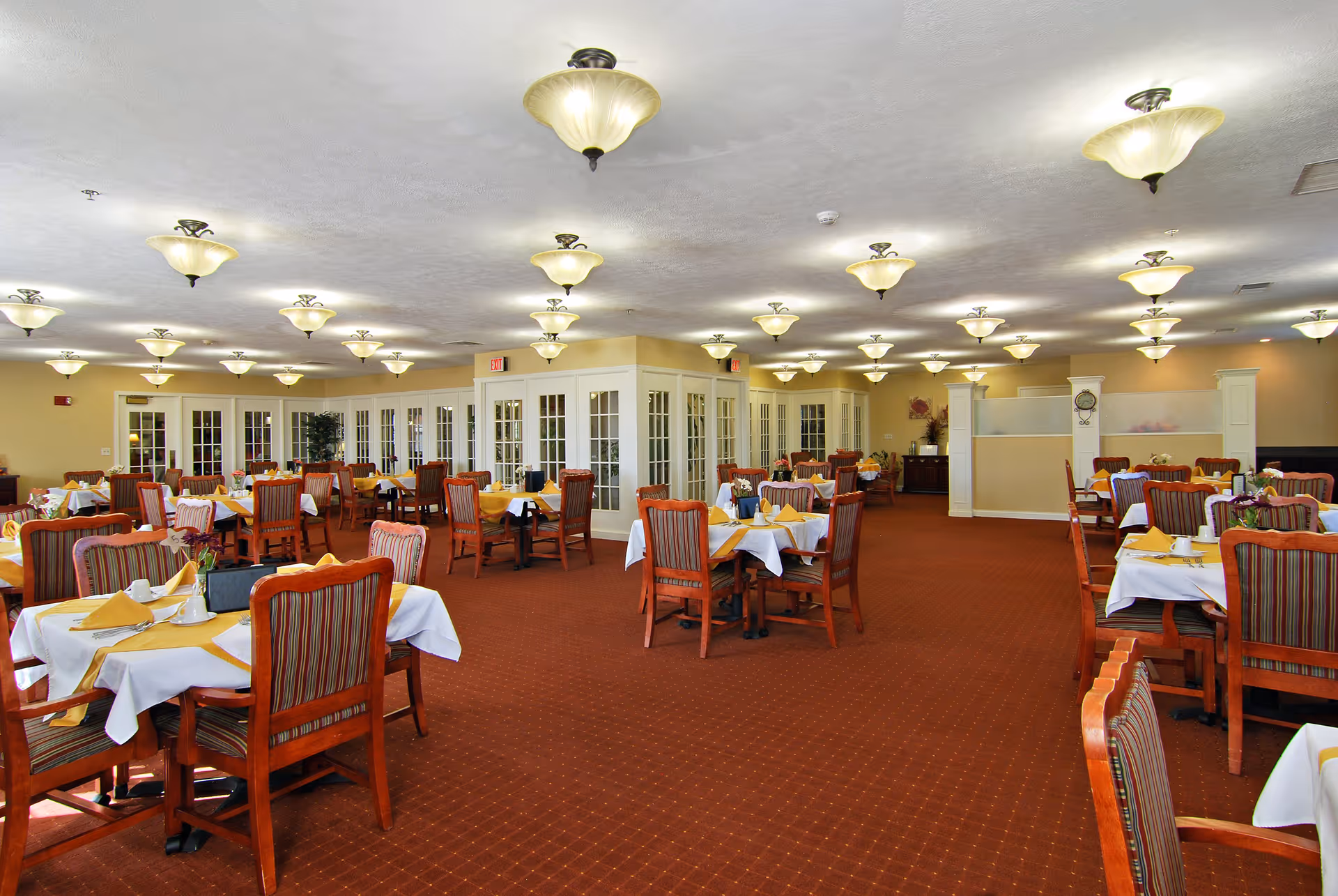 A spacious dining room with multiple tables covered with white tablecloths and yellow napkins. Each table is surrounded by wooden chairs with striped upholstery. The room has a red carpet, cream-colored walls, and numerous ceiling light fixtures. French doors and some plants are visible in the background.
