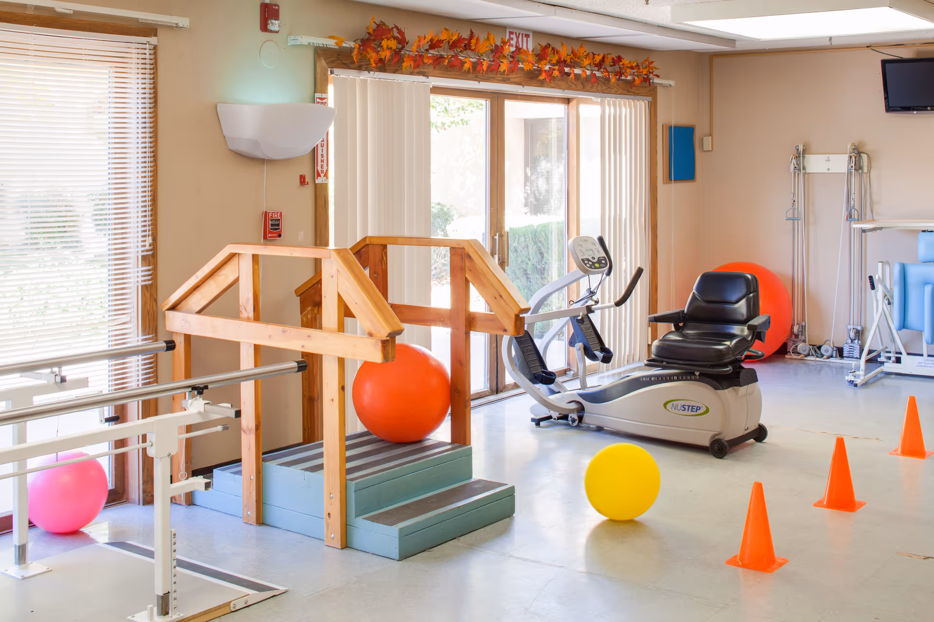 A bright physical therapy room with wooden handrails over small steps, exercise balls in pink, orange, and yellow, an exercise bike, orange cones, and various therapy equipment. Large windows and glass doors let in natural light, and there is a fall-themed garland above the door.
