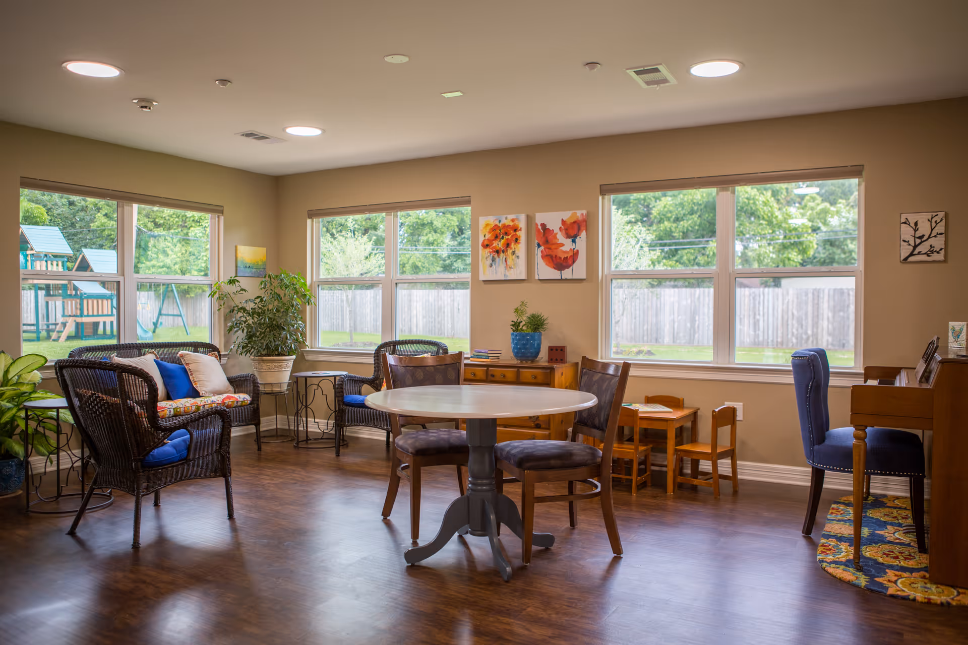 A bright and cozy common area in an assisted living facility with large windows showing a fenced backyard and playground. The room features wicker chairs with cushions, a round table with chairs, a small wooden children's table and chairs, a wooden sideboard with plants and artwork above it, and a piano with a blue cushioned chair on a colorful rug.