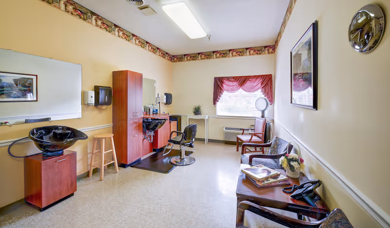 A brightly lit salon room with a hair-washing sink, styling chair, cabinets, and a small waiting area with chairs by a window.