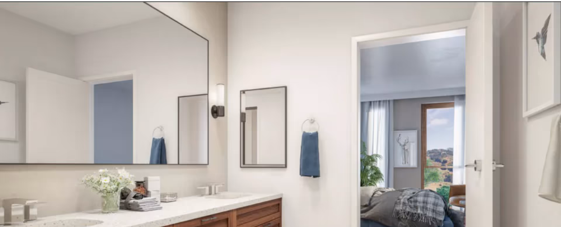 A bright bathroom with a long vanity and large mirror, double sinks and countertop decor, and an open door revealing a bedroom with a bed and window view.