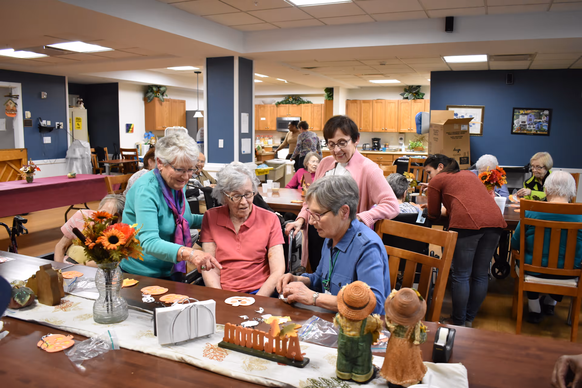 A group of elderly women and caregivers engaging in a craft activity around a large wooden table in a communal room with a kitchen area in the background. The table is decorated with autumn-themed items including small scarecrow figurines and a vase with flowers. The room has a warm and welcoming atmosphere with several people interacting and participating in the activity.
