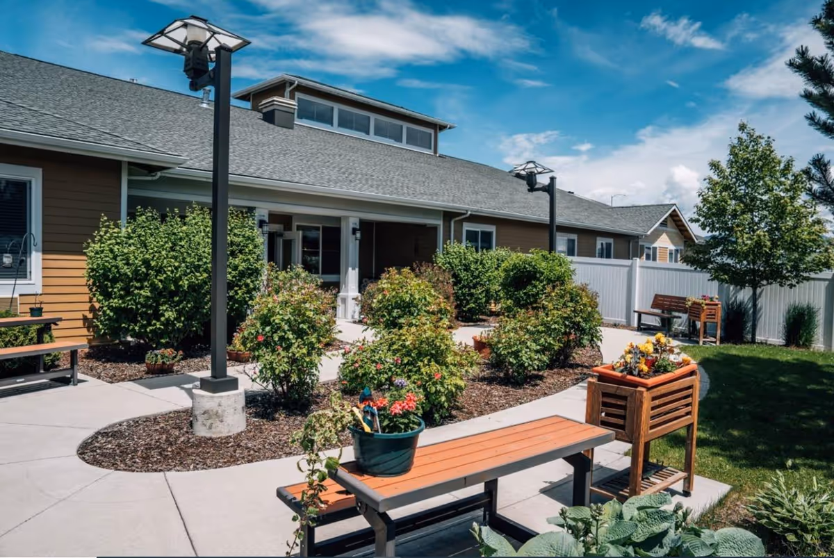 Outdoor garden area at Kalispell Assisted Living featuring a paved walkway, benches, flower pots, bushes, and a tree under a partly cloudy blue sky.