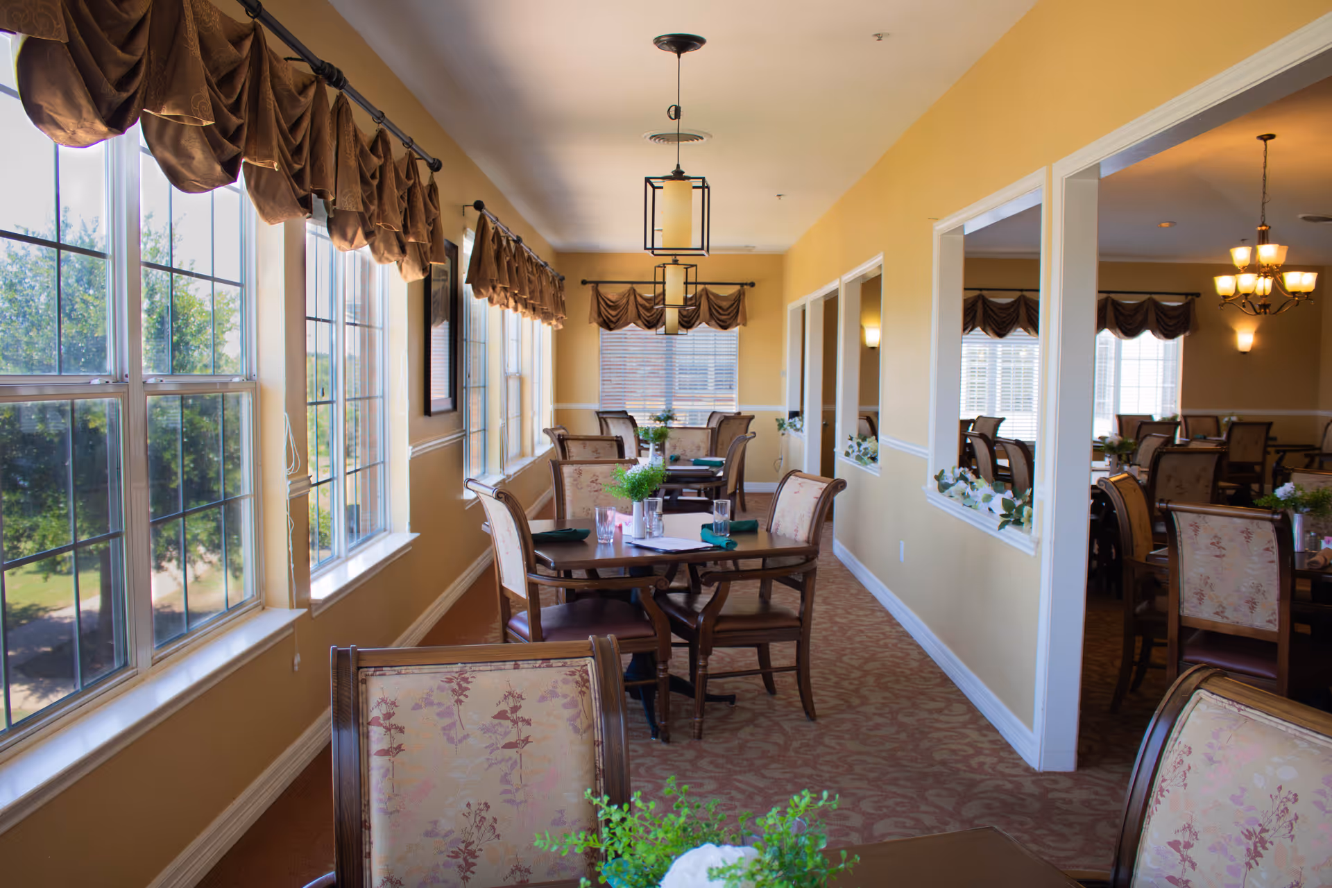 A bright dining room with multiple wooden tables and upholstered chairs arranged neatly. Large windows on the left side allow natural light to fill the room, with brown valance curtains hanging above. The walls are painted a warm yellow, and decorative plants and table settings are placed on the tables. The room extends into another dining area visible through open wall sections.