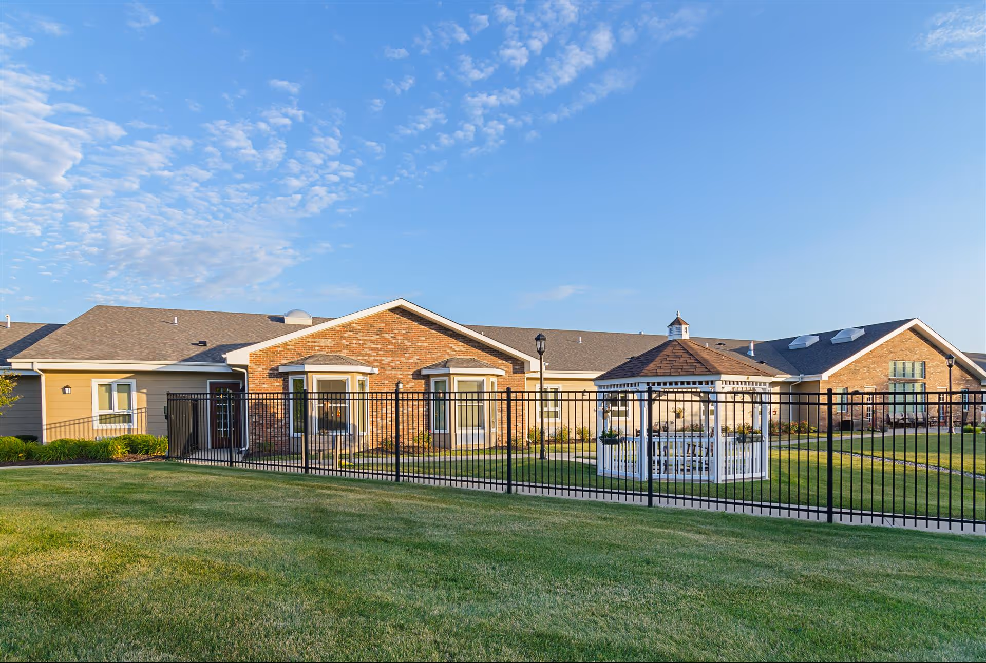 Exterior view of The Gardens at Barry Road Assisted Living and Memory Care facility showing a brick and siding building with multiple windows, a black metal fence, a white gazebo with seating inside, green grass lawn, and a clear blue sky with scattered clouds.