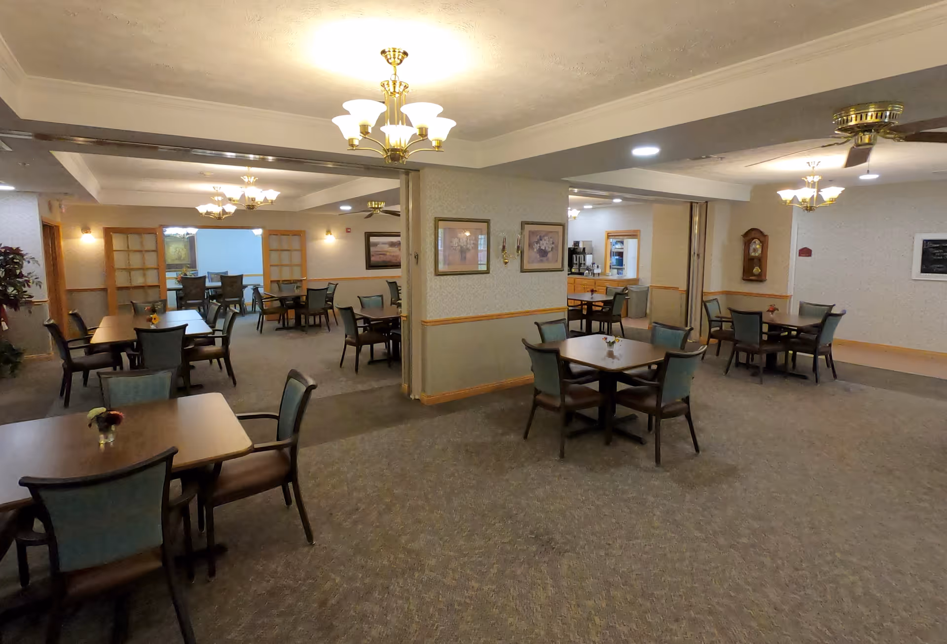 A spacious dining room in an assisted living facility with multiple wooden tables and chairs arranged neatly. The room features beige walls with framed artwork, carpeted floors, and several ceiling light fixtures including chandeliers and ceiling fans. There is a small counter area in the background with a coffee machine and additional seating.