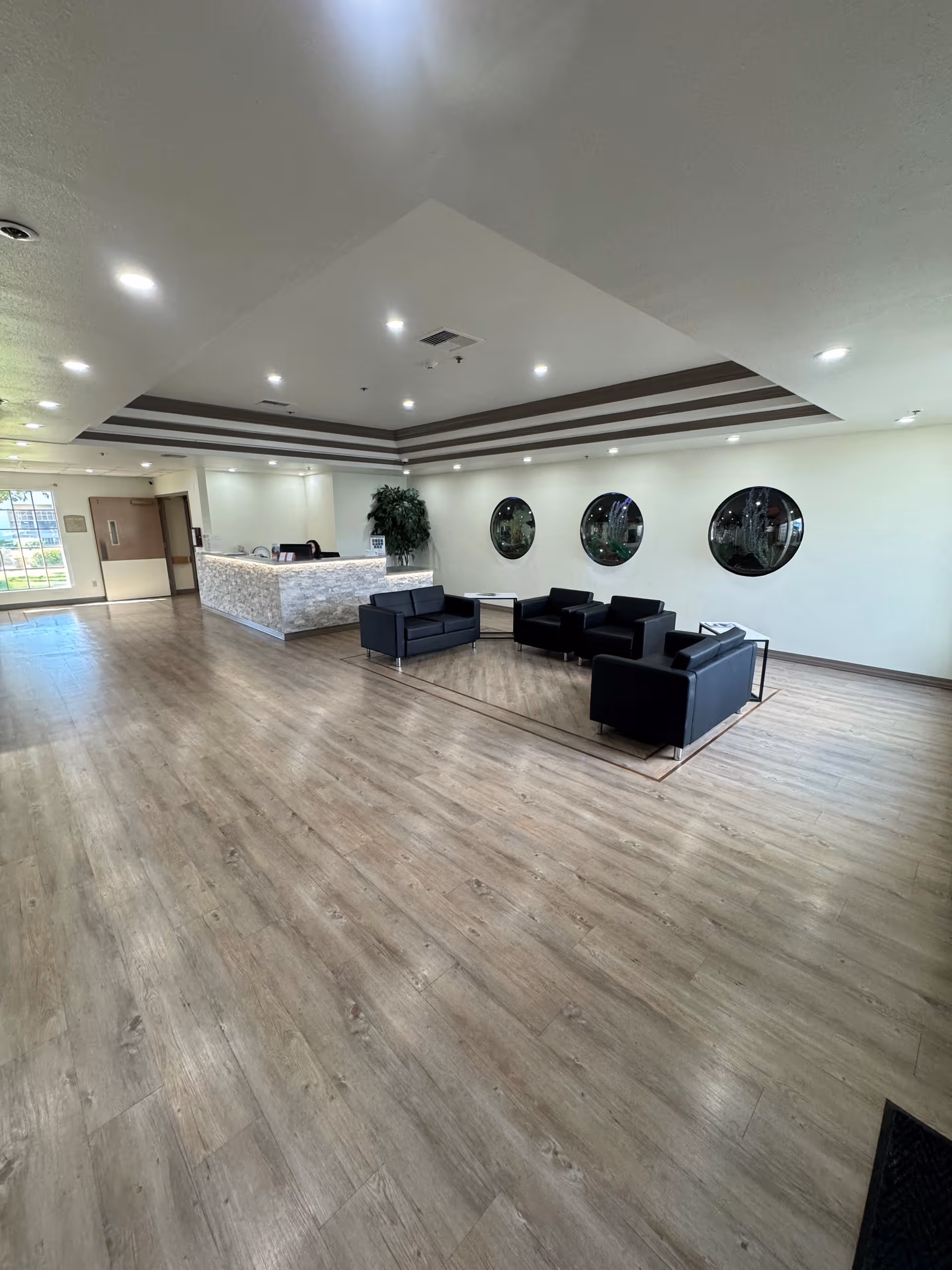 A spacious and modern lobby area with light wood flooring, a white stone reception desk, and a seating area consisting of black leather sofas and armchairs arranged around a small table. The walls are white with three round windows, and there is a large plant near the reception desk. The ceiling has recessed lighting and decorative trim.