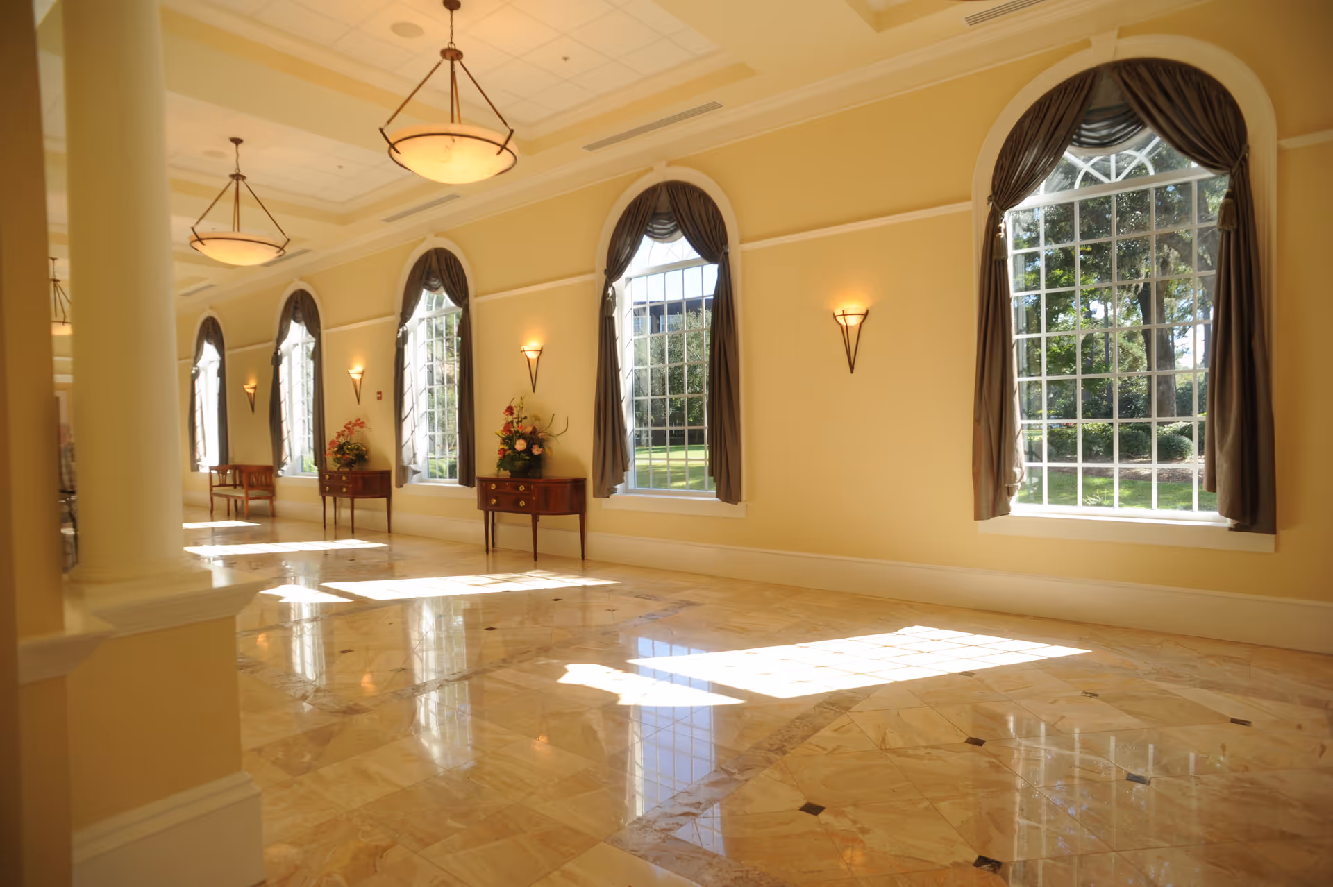 Bright, elegant lobby corridor with arched windows, marble floor, chandeliers and small tables with floral arrangements.