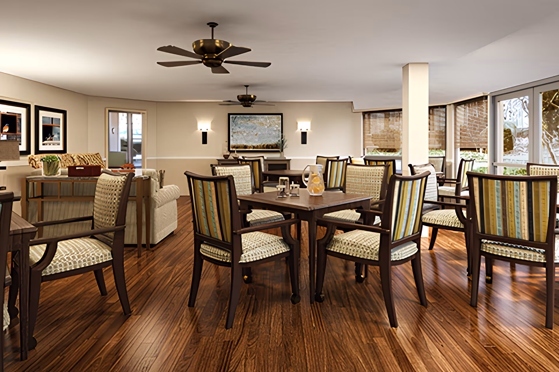 A spacious and well-lit common area in a senior living facility featuring multiple wooden tables surrounded by cushioned chairs with striped upholstery. The room has wooden flooring, ceiling fans, wall sconces, framed artwork, and large windows with blinds allowing natural light to enter. There are also some comfortable armchairs and a console table with decorative items.