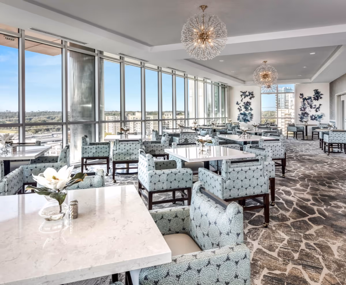Bright and spacious dining room with large floor-to-ceiling windows offering a city view. The room features multiple square tables with patterned upholstered chairs, decorative flower arrangements on each table, modern chandeliers hanging from the ceiling, and artistic wall decor.