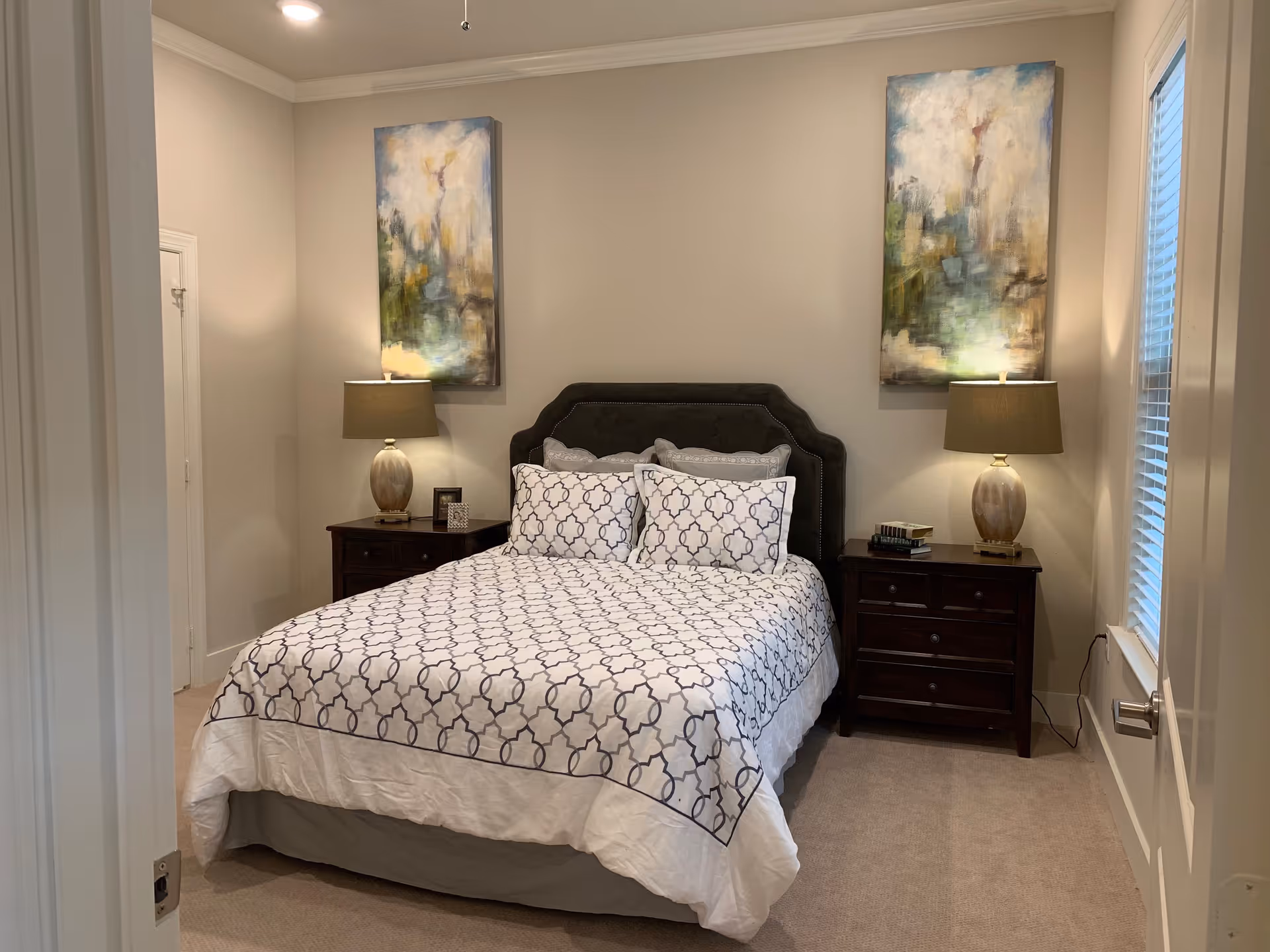A neatly made bed with a patterned white and gray bedspread and matching pillows in a bedroom. The bed has a dark upholstered headboard. On each side of the bed are dark wooden nightstands with table lamps and decorative items. Two abstract paintings hang on the wall above the nightstands. A window with blinds is visible on the right side of the room.