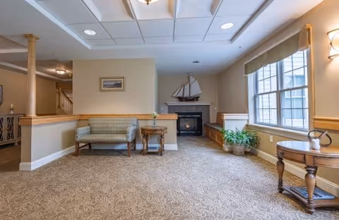 Bright common room with upholstered seating, side tables, a fireplace topped by a model sailboat, and a large window.