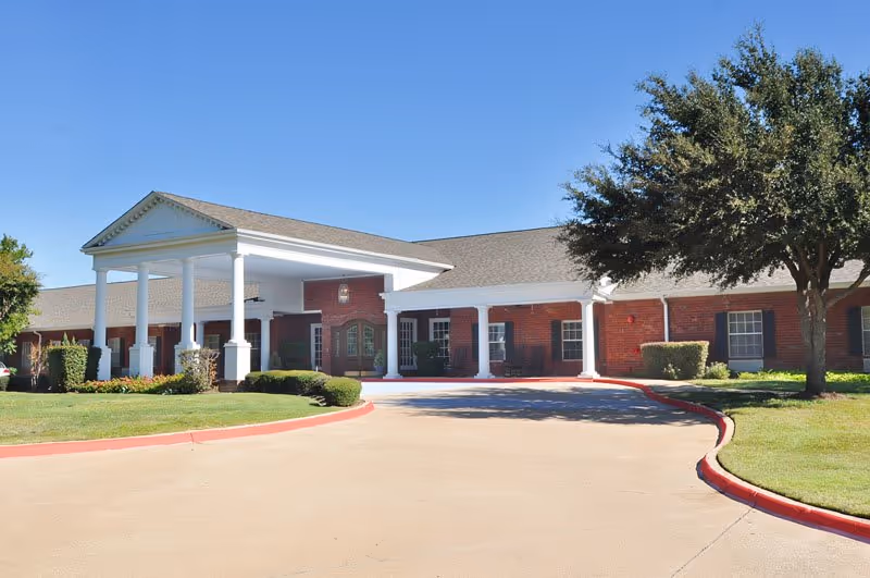 Exterior view of a single-story senior living facility building with a covered entrance supported by white columns, red brick walls, and a curved driveway surrounded by green grass and trees under a clear blue sky.